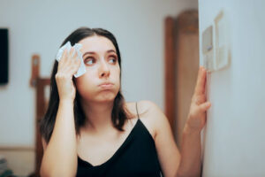 Woman wiping her sweating forehead while looking at her thermostat settings.