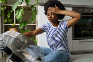 Woman holding hand to her sweating forehead as she sits in front of fan due to broken AC.